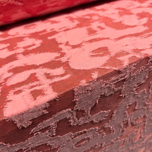 May include: Close-up of a pink fabric with a textured, embossed pattern. The fabric has a subtle sheen and a slightly rough texture.