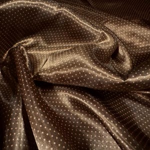 May include: Close-up of a brown satin fabric with a subtle sheen. The fabric is covered in small, evenly spaced white polka dots. The material is draped, creating soft folds and shadows, highlighting its texture and sheen.