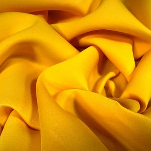 May include: Close-up of a vibrant yellow fabric with soft folds and a silky texture. The material is tightly bunched, creating a dynamic interplay of light and shadow. The fabric's color is a rich, sunny yellow.