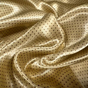 May include: A close-up of a gold satin fabric with small brown polka dots. The fabric is draped and folded, showing its texture and sheen.