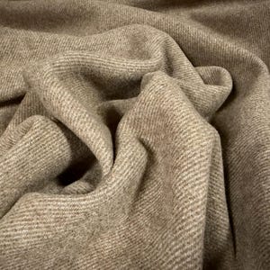 May include: A light brown fabric with a visible diagonal twill weave pattern. The material is folded, creating soft, deep ripples and shadows that highlight the textured surface and the consistent, neutral tan color throughout the textile.