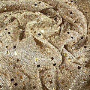 May include: Close-up of a shimmering gold fabric with a textured weave. The fabric is adorned with small, square, gold sequins scattered across its surface. The material is draped, creating soft folds and shadows.