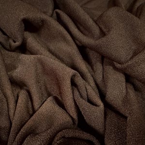 May include: Close-up of a dark brown fabric with a textured surface. The fabric is draped and folded, creating shadows and highlights. The material appears to be soft and flowing, suitable for clothing or home decor.