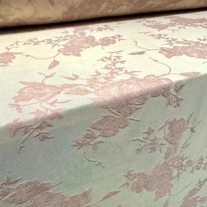 May include: Close-up of a fabric roll with a floral design. The fabric has a light beige background with a raised, textured design of pink flowers and foliage. The pattern is repeated across the material.