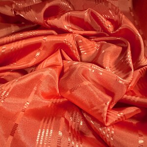 May include: Close-up of a vibrant, coral-colored fabric with a subtle sheen. The material is draped and folded, creating shadows and highlights. A repeating pattern of small, rectangular, light-colored accents adds texture to the fabric.