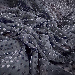 May include: Dark grey fabric featuring a pattern of small, evenly spaced, white and black polka dots. The fabric is draped, creating soft folds and shadows. The material appears lightweight and flowing, suitable for clothing or decorative projects.