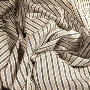 May include: Close-up of a fabric with a striped pattern. The fabric features vertical stripes in shades of beige, gray, and white. The fabric is draped, creating folds and shadows that add depth and texture to the image.