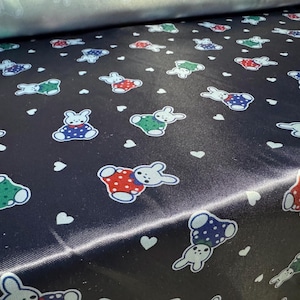 May include: Black satin fabric with a repeating pattern of white hearts and colorful cartoon bunnies. The bunnies are wearing different colored clothes and have white accents.