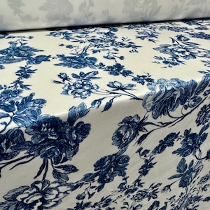 May include: Off-white fabric with a repeating pattern of blue floral designs. The pattern features detailed illustrations of roses and other flowers, creating a classic, elegant aesthetic. The fabric is likely used for upholstery or home decor.