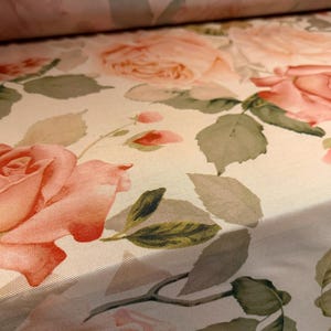 May include: Close-up of a fabric featuring a floral design. Large, soft pink roses and green leaves are set against a cream background. The fabric is rolled, revealing the pattern's repeat.