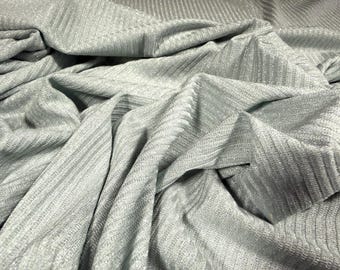 May include: Close-up of a light gray fabric with a ribbed texture. The fabric is draped and folded, creating shadows and highlights. The material has a subtle sheen, suggesting a blend of fibers. The overall appearance is soft and flowing.