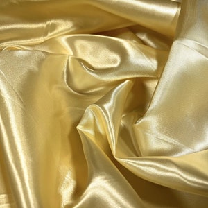 May include: A close-up of a smooth, silky fabric in a pale yellow color. The fabric is draped and folded, creating soft, flowing lines.