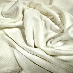 May include: A close-up of a white fabric with a soft, smooth texture. The fabric is draped and folded, creating a sense of movement and flow.