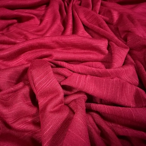 May include: Close-up of a deep red fabric with a textured weave. The fabric is draped and folded, creating shadows and highlights that emphasize its texture and color. The material appears soft and flowing.