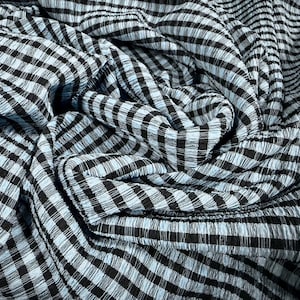Könnte beinhalten: Nahaufnahme eines Stoffes mit einem schwarz-hellblauen Karomuster. Der Stoff ist strukturiert und scheint drapiert zu sein, mit sichtbaren Falten und Knitterfalten. Das Muster ist ein klassisches Vichy-Design.