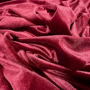 Könnte beinhalten: Nahaufnahme eines tiefroten Samtstoffs mit weichen Falten und einer strukturierten Oberfläche. Der Stoff hat ein sattes, luxuriöses Aussehen, mit Lichtreflexen und Schatten, die seine Tiefe und Textur hervorheben. Der Gesamteindruck ist von Opulenz und Komfort geprägt.