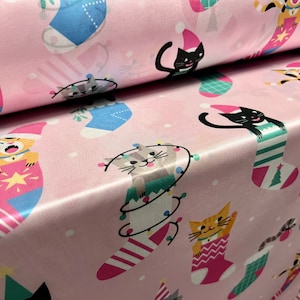 May include: Pink fabric with a repeating pattern of cats wearing Christmas stockings. The cats are in various poses and have different colored stockings. The background is a light pink with white snowflakes.
