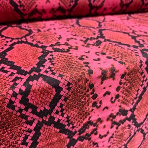May include: A close-up shot of a pink fabric with a snakeskin design. The pattern features scales in shades of pink, red, and brown, with black outlines. The fabric is folded, revealing its texture and the depth of the material.