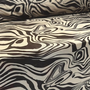 May include: Close-up of a fabric with a zebra stripe pattern. The fabric is a cream color with black stripes that curve and swirl across the surface. The fabric is folded over in the upper left corner.