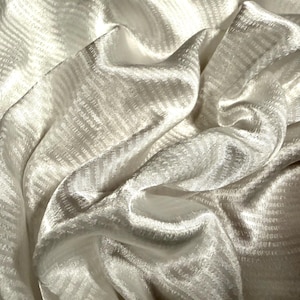 May include: Close-up of a white fabric with a subtle, textured pattern. The fabric is draped, creating soft folds and shadows. The material appears to be silky or satin-like, reflecting light and adding depth to the image.