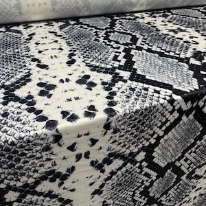 Puede incluir: Primer plano de una tela con estampado de piel de serpiente. El diseño presenta un patrón repetitivo de escamas negras, blancas y grises, creando un efecto realista de piel de reptil. La tela está doblada, mostrando la textura y la caída del material.
