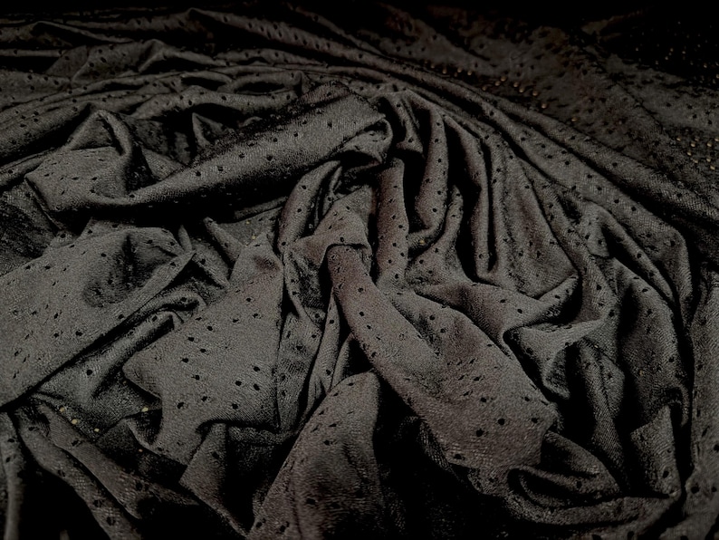 May include: A dark black fabric featuring a consistent pattern of small, evenly spaced circular perforations throughout the material. The textile is draped in deep folds, creating a textured surface with varying shadows and highlights.
