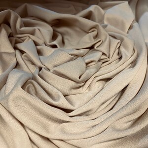 Könnte beinhalten: Nahaufnahme eines beigefarbenen Stoffes, kunstvoll drapiert und gefaltet. Der Stoff hat eine subtile Textur, mit weichen Falten und Schatten, die Tiefe erzeugen. Die neutrale Farbe deutet auf Vielseitigkeit für verschiedene Näh- und Bastelprojekte hin.