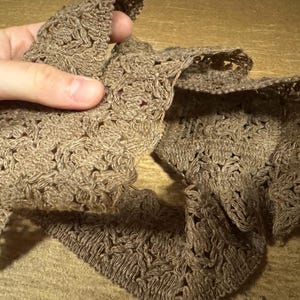 May include: Close-up of a length of brown, open-weave trim. The trim is held in a hand, showing the intricate floral pattern. The trim appears to be made of a natural fibre, possibly linen or cotton, and has a slightly textured surface.