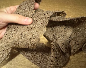May include: Close-up of a length of brown, open-weave trim. The trim is held in a hand, showing the intricate floral pattern. The trim appears to be made of a natural fiber, possibly linen or cotton, and has a slightly textured surface.