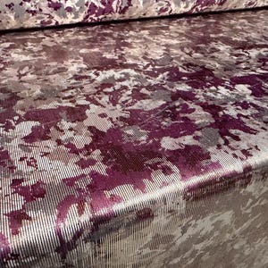 May include: Close-up of a fabric with a textured surface. The fabric features a pattern of purple and gray brushstrokes over a silver background. The fabric is folded, revealing the pattern's repeat.