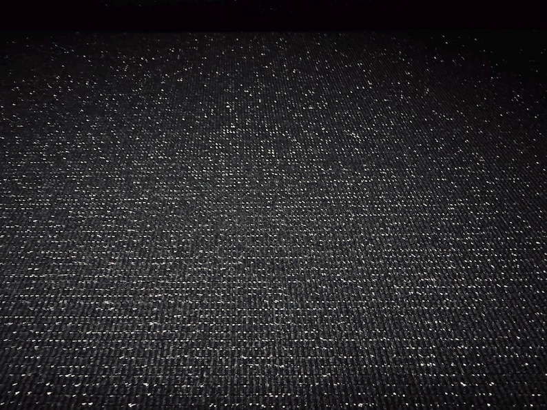 May include: A close-up view of a black fabric with a subtle, textured weave. The fabric is densely covered with tiny, sparkling silver flecks, creating a shimmering effect. The overall appearance is dark and elegant.