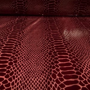 Puede incluir: Primer plano de un patr&oacute;n de piel de serpiente burdeos. El material de cuero sint&eacute;tico tiene un acabado brillante y un dise&ntilde;o de escamas repetitivo. La tela est&aacute; doblada en la parte superior, revelando la parte inferior. El color es rojo intenso.