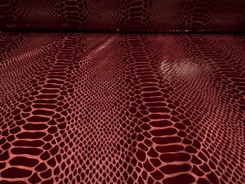 Puede incluir: Primer plano de un patr&oacute;n de piel de serpiente burdeos. El material de cuero sint&eacute;tico tiene un acabado brillante y un dise&ntilde;o de escamas repetitivo. La tela est&aacute; doblada en la parte superior, revelando la parte inferior. El color es rojo intenso.
