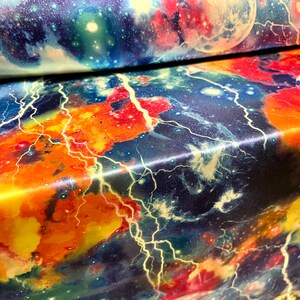 May include: A colorful fabric with a galaxy print featuring lightning bolts, stars, and planets. The fabric has a shiny, satin-like finish.