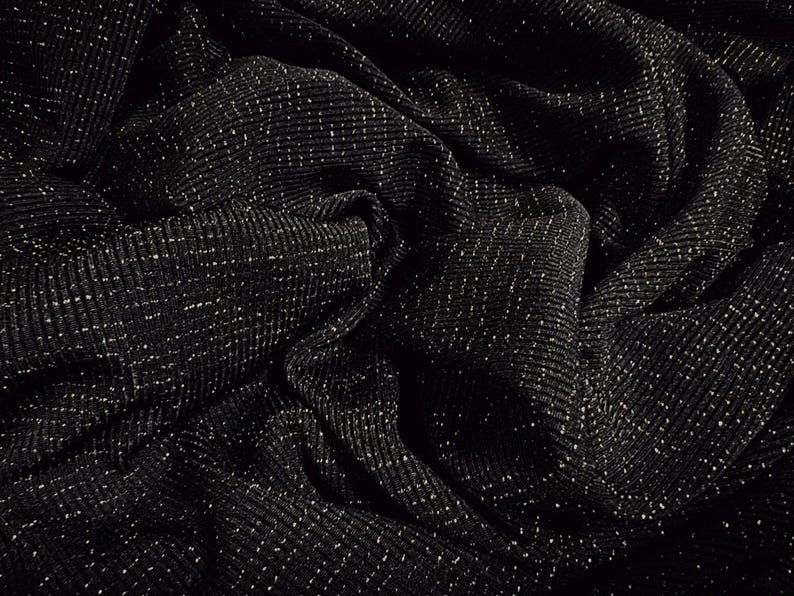 May include: Close-up of a black fabric with a ribbed texture and a subtle sparkle. The material is draped, creating folds and shadows that enhance the visual depth. The fabric's surface is dotted with tiny, reflective specks.