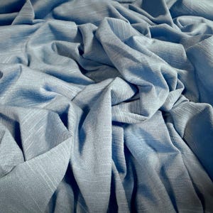 May include: Close-up of a pale blue fabric with a textured weave. The fabric is draped and folded, creating shadows and highlights. The material appears soft and flowing, suitable for clothing or home decor.