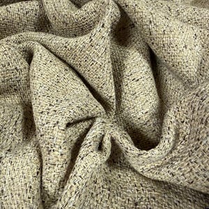 Könnte beinhalten: Ein strukturierter Stoff mit einem Webmuster aus hellbeigen und hellbraunen Fäden, durchsetzt mit kleinen schwarzen Sprenkeln. Das Material ist in weichen Falten drapiert, was die grobe, ineinandergreifende Struktur des Gewebes hervorhebt.
