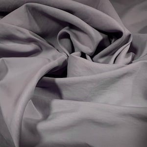 Puede incluir: Primer plano de una tela suave y gris con una textura lisa. La tela está drapeada y doblada, creando una serie de pliegues y curvas.