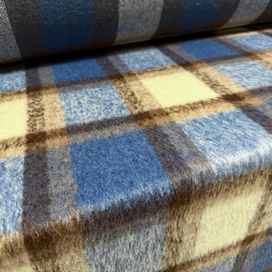 May include: A bolt of fabric featuring a plaid pattern with a soft, fuzzy texture. The design consists of intersecting lines in shades of blue, cream, and dark brown, creating a repeating square grid across the surface of the material.