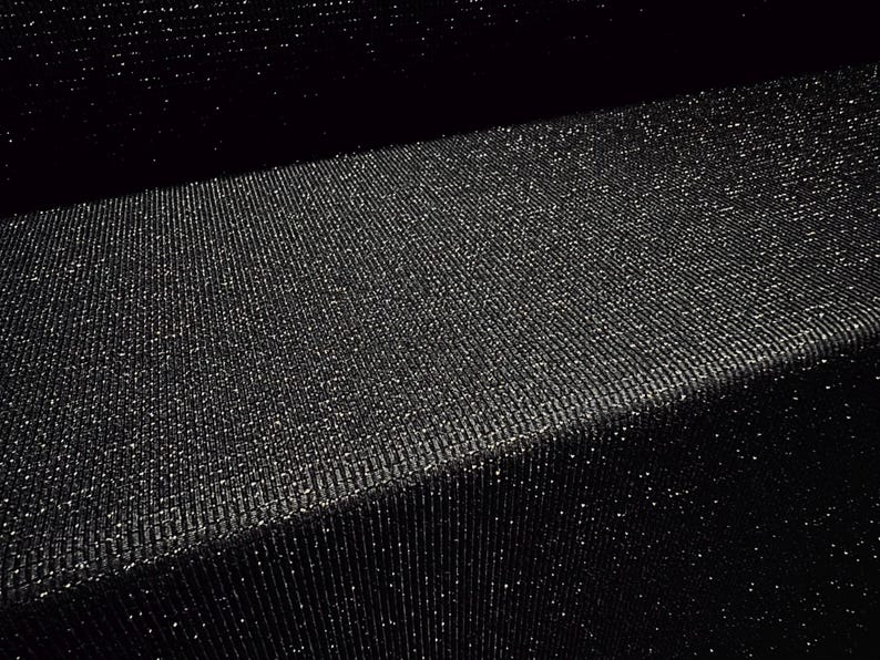 May include: Close-up of a black fabric with a textured, ribbed surface. The fabric has a subtle sparkle, catching the light and creating a shimmering effect. The material is folded, showing depth and dimension.