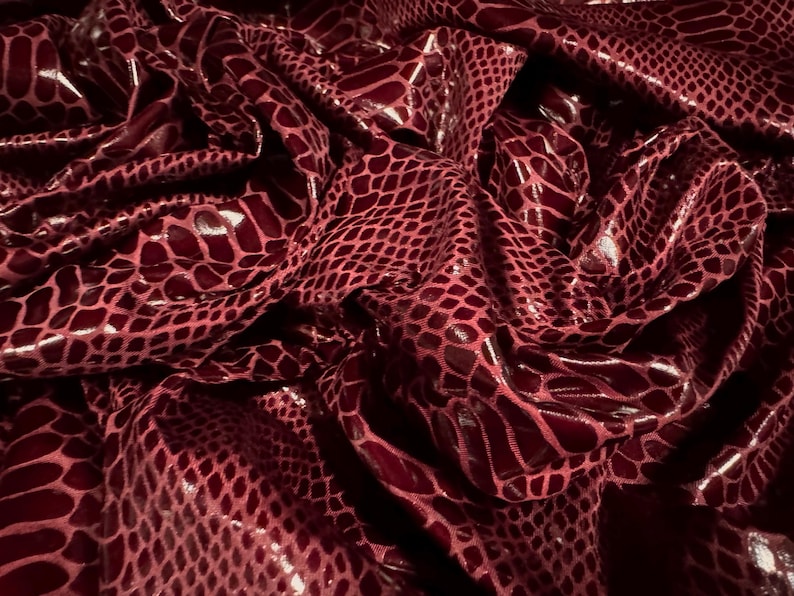 Puede incluir: Primer plano de una tela burdeos con una superficie brillante y texturizada. La tela presenta un patr&oacute;n de escamas de reptil. La tela est&aacute; arrugada y doblada, creando sombras y reflejos.
