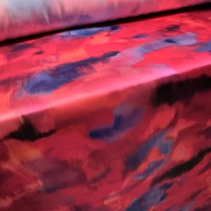 May include: Red fabric with a watercolor-like pattern of blue, pink, and black. The fabric has a smooth, silky texture.