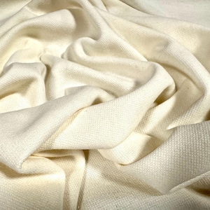 May include: Close-up of a cream-coloured fabric with a textured weave. The fabric is draped and folded, creating soft shadows and highlights. The material appears to be a medium-weight textile, suitable for various sewing projects.