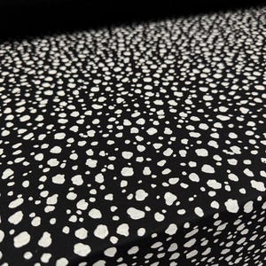 Pode incluir: Tecido preto com um padrão de manchas brancas. O tecido apresenta um design repetitivo de formas brancas irregulares sobre um fundo preto. Adequado para roupas ou decoração de casa.