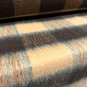 May include: A thick, fuzzy fabric featuring a large-scale plaid pattern in shades of beige, dark brown, and soft light blue. The material has a visible, soft-textured nap and is folded in layers, showcasing the repeating checkered design throughout.