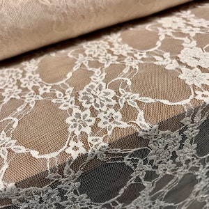May include: Close-up of white floral lace fabric. The delicate lace features intricate floral patterns and a fine mesh backing. A rolled section of the fabric is visible in the background, showcasing the texture and detail of the material.