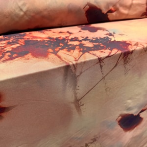 May include: Close-up of a beige fabric with a brown and black abstract print. The print resembles a tree branch with leaves and has a faded, watercolour effect.