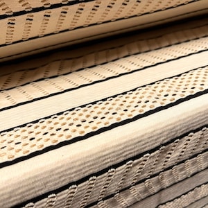 Peut inclure: Gros plan sur un tissu crème à motifs rayés. Le motif présente des bandes alternées de crème unie, de noir et de sections perforées avec de petits trous espacés. Le tissu est plié, montrant plusieurs couches.