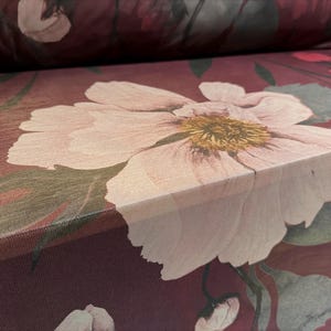 May include: Close-up of a floral fabric featuring a large, pale pink flower with a yellow centre. The fabric has a deep burgundy background with green leaves and other floral elements. Suitable for upholstery or home decor.
