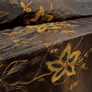 Op de afbeelding: Bruin stof met een gouden geborduurd bloemenpatroon. De stof heeft een gekreukelde textuur.